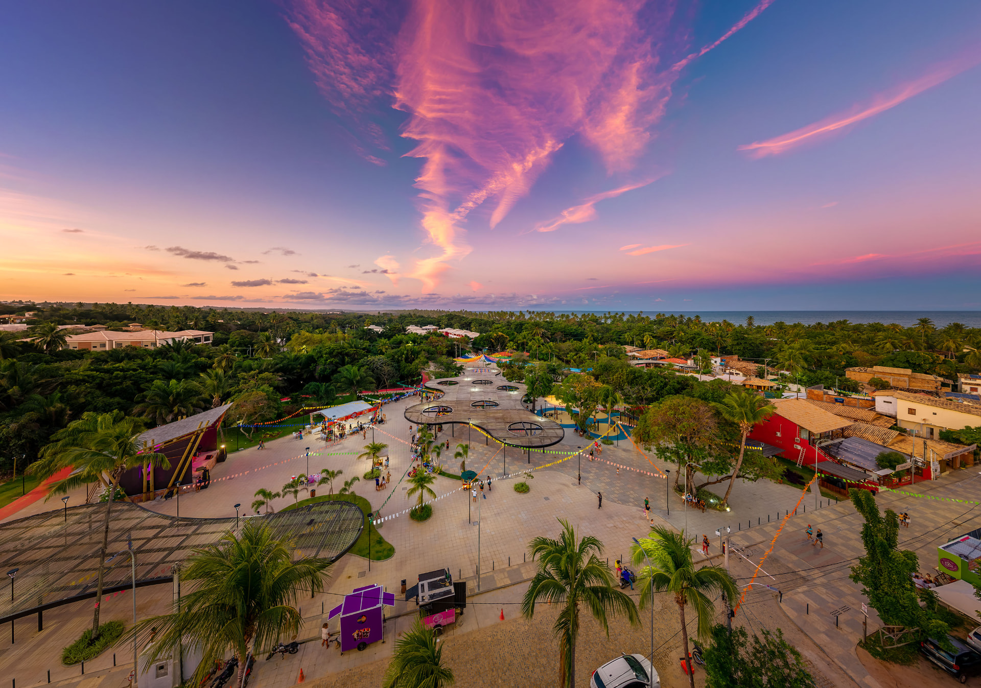 Vista aérea da vila reformada de Imbassaí ao pôr do sol, com praça de piso claro, coberturas de palha, vegetação nativa e o mar ao fundo